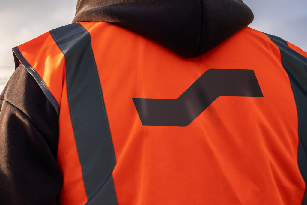 A man with a orange high-vis on, facing away from the camera, the camera is focused on the SYPN on the back of the high-vis.